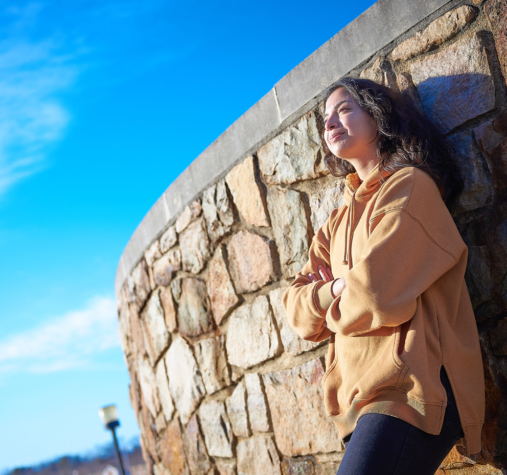 A female student leaning on a wall against a bright blue sky